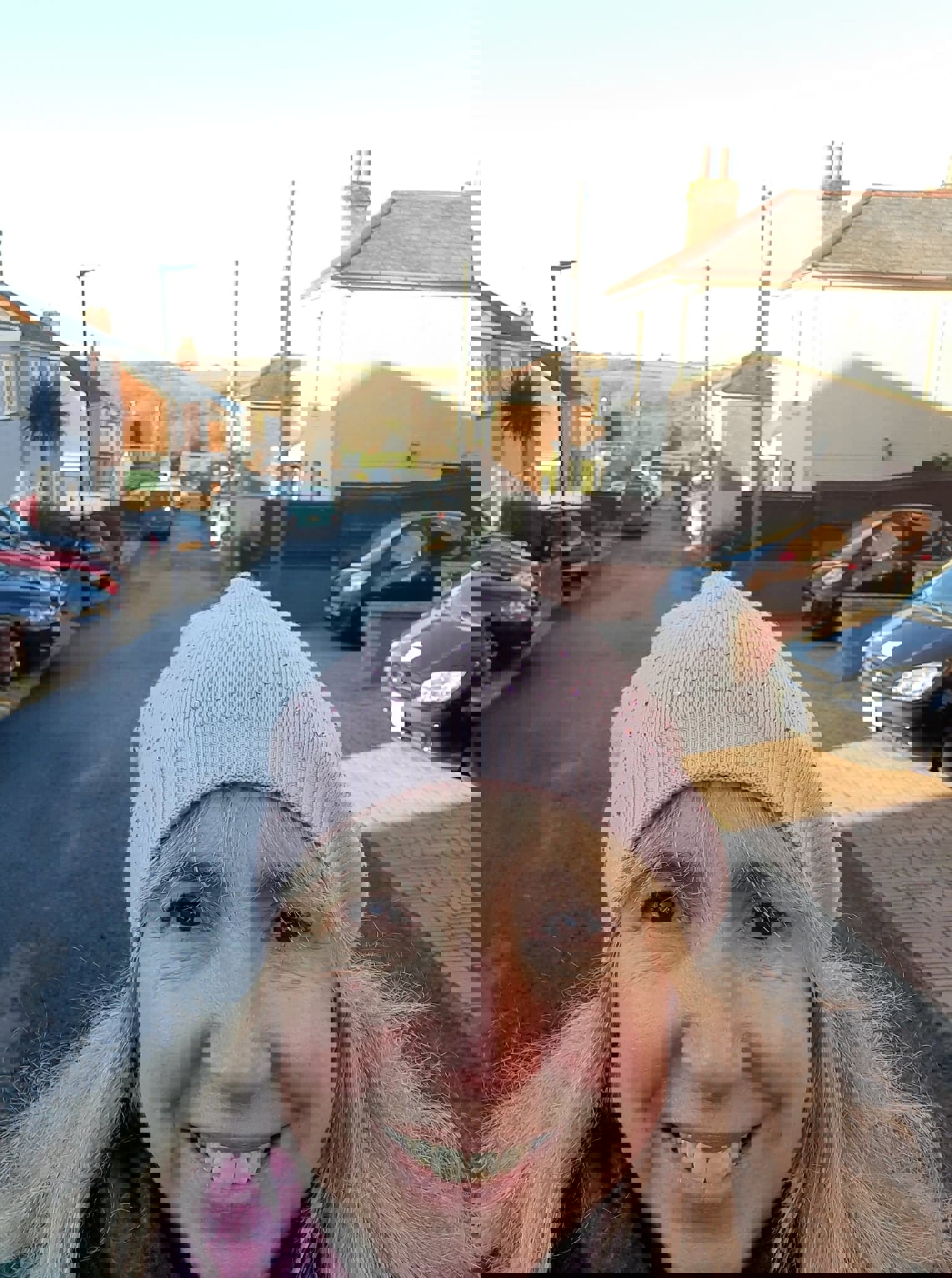 woman with long blonde hair wearing pink woolly hat with residential street behind her